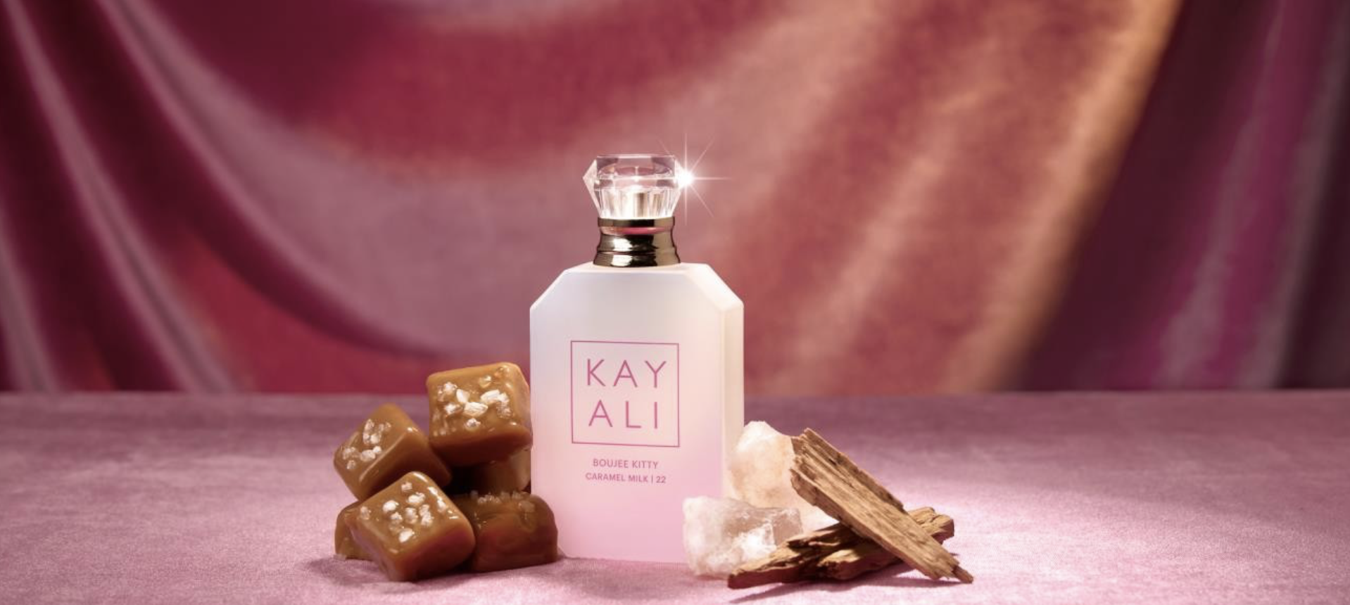 KAYALI Boujee Kitty Caramel Milk | 22 Eau de Parfum bottle beside salted caramel cubes and sandalwood on pink velvet